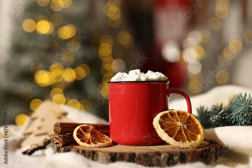 Cup of cocoa with marshmallows, cinnamon sticks, dry orange slices and Christmas decor on table against festive lights, closeup