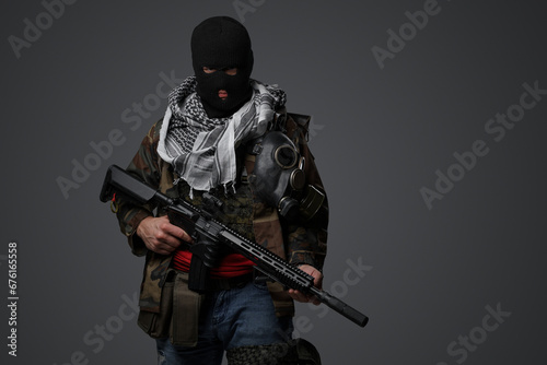 A Middle Eastern radical soldier dressed in a black balaclava and camouflaged field uniform, armed with a rifle, against a gray background photo