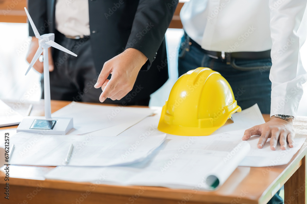 A meeting of two male engineers in an office is pointing at a wind turbine model regarding the future design of the energy industry. On the table are hard hats, blueprints, pens. A meeting of two male engineers in an office is pointing at a wind turbine model regarding the future design of the energy industry. On the table are hard hats, blueprints, pens.