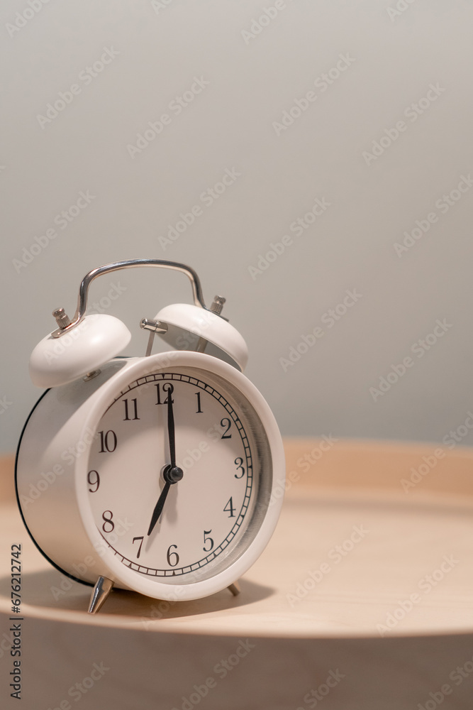 Close-up of a round white alarm clock on a table in the bedroom. The hands on the clock show seven o'clock in the morning, time to get up. Retro alarm clock on the table, vintage tone. space for text Close-up of a round white alarm clock on a table in the bedroom. The hands on the clock show seven o'clock in the morning, time to get up. Retro alarm clock on the table, vintage tone. space for text