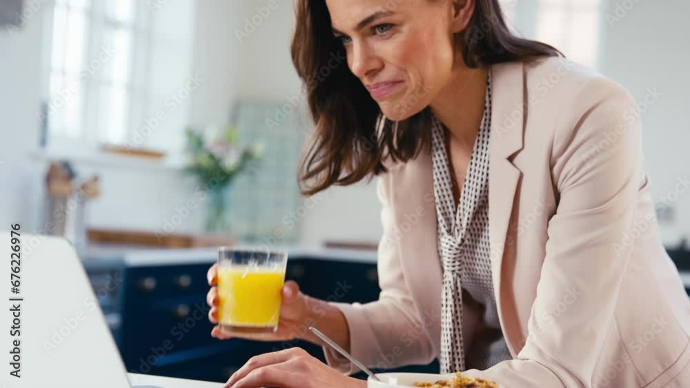 Businesswoman at home multi-tasking in kitchen working on laptop whilst eating breakfast and drinking orange juice - shot in slow motion Businesswoman at home multi-tasking in kitchen working on laptop whilst eating breakfast and drinking orange juice - shot in slow motion