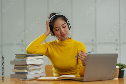 Confident Asian female student wearing wireless headphones studying online, searching for information, thinking analytically from documents, textbooks, laptop computer for business studies. photo