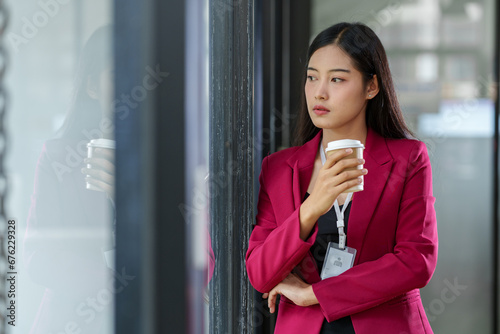 Young Asian accountant or businesswoman with a positive attitude stands holding a paper coffee cup, thinking, absentmindedly, about work, planning, company management in the office.
