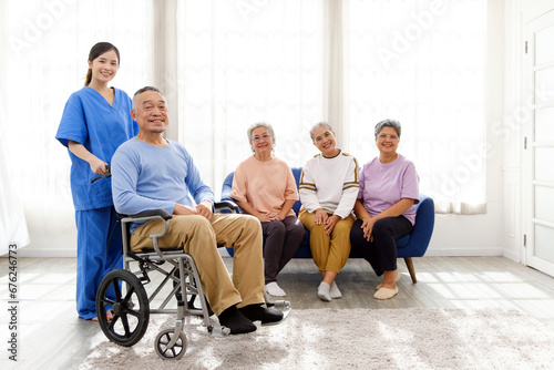 The caregiver therapist stands with an Asian senior sitting in a wheelchair with a group of senior women sitting on a sofa in the background. The nursing home facilitates a support group.