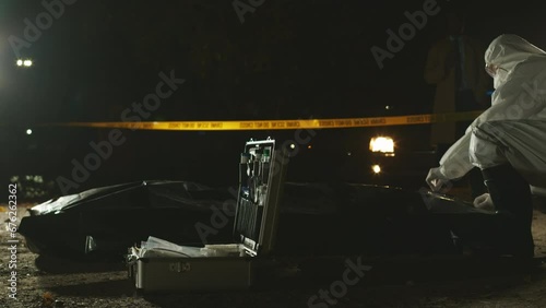 Unrecognizable forensic specialist in protective coveralls opening black plastic bag to examine corpse lying on ground at night. Policeman standing near car with flashing lights in dark backgroundUnre photo