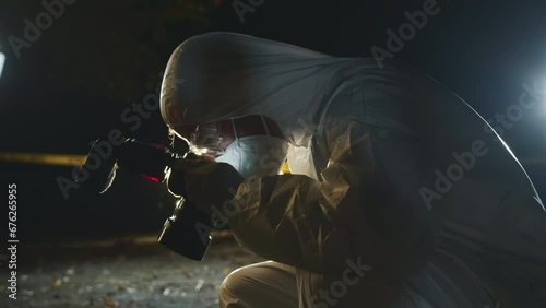 Tilt up of unrecognizable forensic expert photographing murder scene at night on professional camera with bright headlights from police car in dark background photo