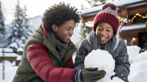 Madre e hijo afroamericanos abrigados jugando con una bola de nieve un día nevado de invierno en la ciudad. photo