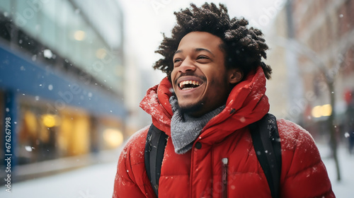 Hombre afroamericano sonriendo de pie con un abrigo rojo y mochila en medio de una calle de la ciudad en un día que esta nevando. photo