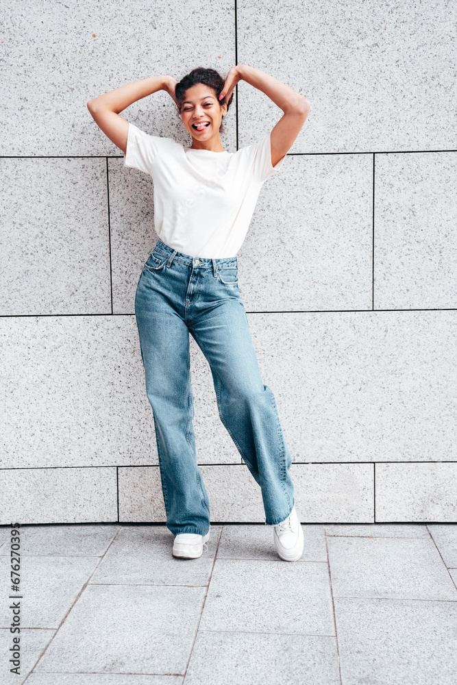 Young beautiful smiling hipster woman in trendy summer white t-shirt and jeans clothes. Carefree woman, posing in the street at sunny day. Positive model outdoors near wall. Going crazy Young beautiful smiling hipster woman in trendy summer white t-shirt and jeans clothes. Carefree woman, posing in the street at sunny day. Positive model outdoors near wall. Going crazy