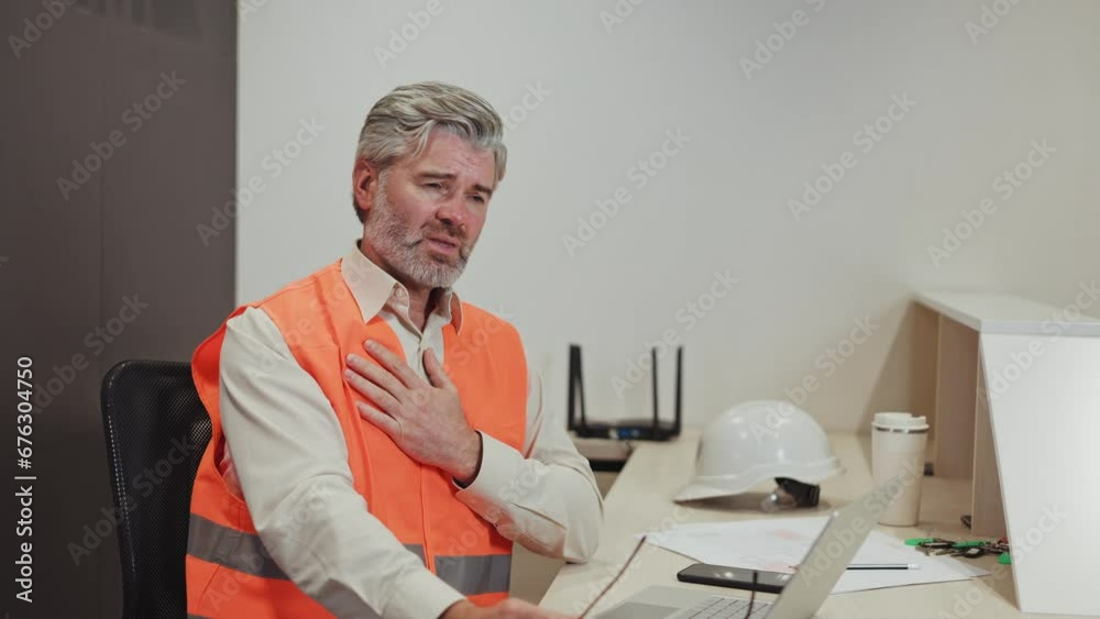 Aged caucasian man suffering from heart disease while sitting at office desk with laptop and smartphone. Exhausted male plant worker in orange vest closing eyes because of severe pain in chest.