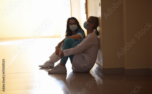 Tired women doctors are sitting in masks in corridor. Burnout of healthcare workers concept