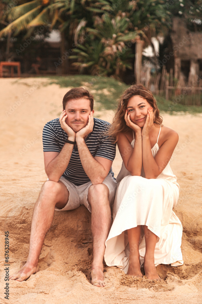 Lovely funny bored couple sitting on sandy beach at tropical ocean, wait looking at camera. Man and woman waiting in travel holiday, coastline. Summer vacation lifestyle concept. Copy ad text space