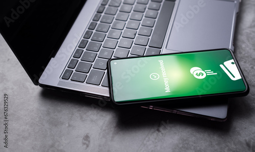 Smartphone laying on laptop with money received notification on screen. Selective focus. Concept of financial transactions with mobile devices. Smartphone screen displaying money transfer confirmation