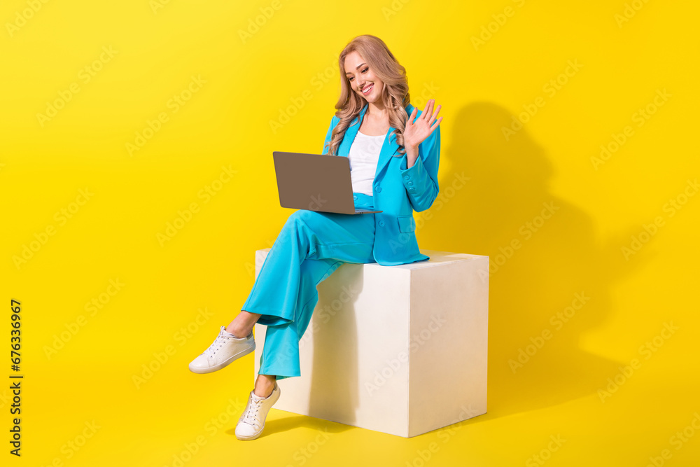 Full length photo of pleasant woman wear blue stylish suit sit on white cube talk on laptop video call isolated on yellow background