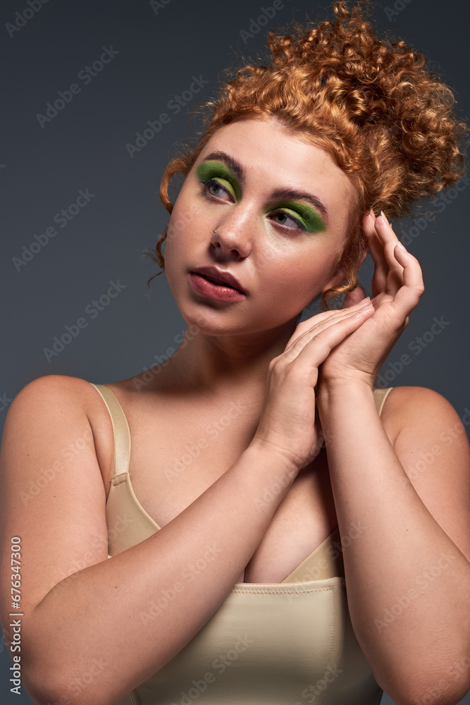 portrait of dreamy curvy woman with red hair and bold makeup looking away on dark grey backdrop