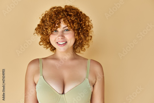 young curvy woman with red wavy hair posing in bra and smiling at camera on beige backdrop