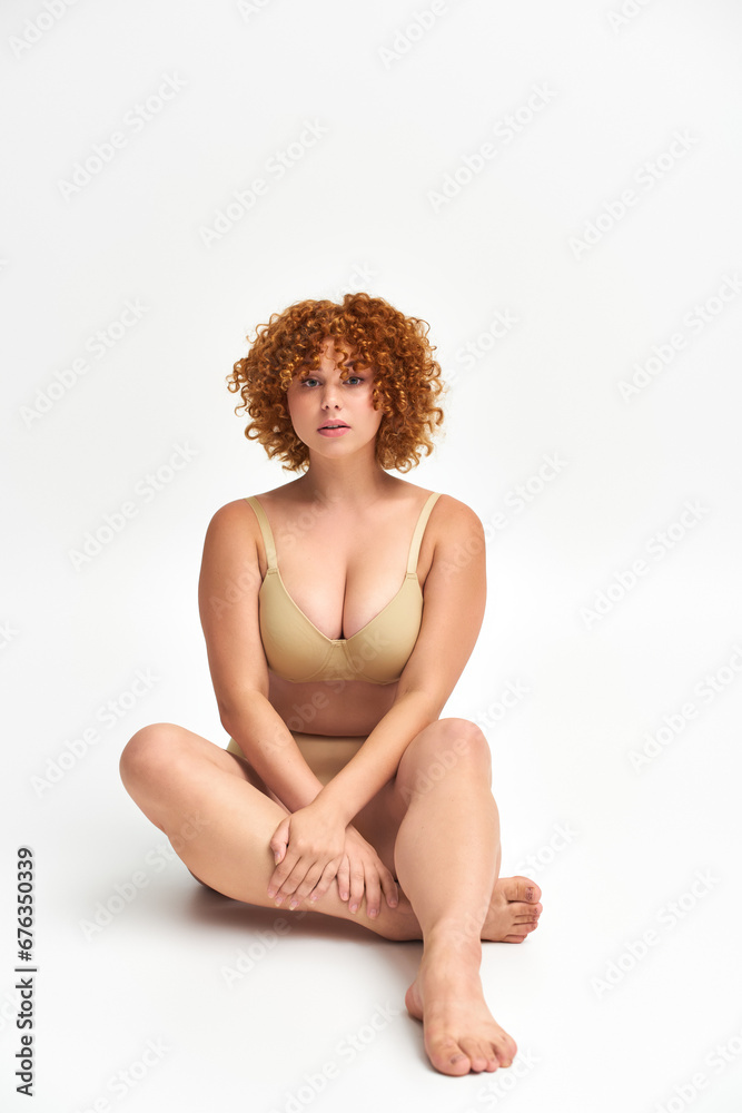 Obraz premium full length of curvaceous redhead woman in beige lingerie sitting and looking at camera on white