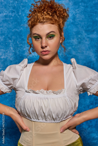 beautiful redhead plus size model in trendy attire with hands on hips on blue textured backdrop
