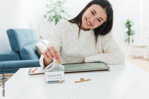 Smiling crop woman sitting with tablet at table and putting coins in house model savings bank