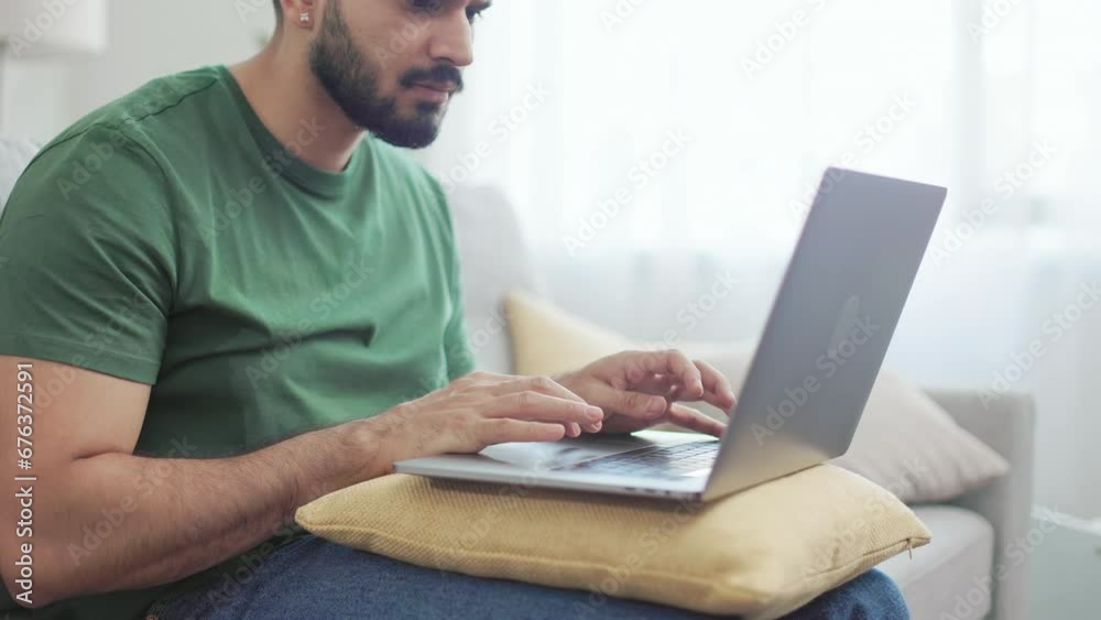 Focused indian freelancer sitting on comfy couch and typing on wireless laptop. Bearded guy in casual wear using yellow soft pillow on knees as support for portable computer. Focused indian freelancer sitting on comfy couch and typing on wireless laptop. Bearded guy in casual wear using yellow soft pillow on knees as support for portable computer.