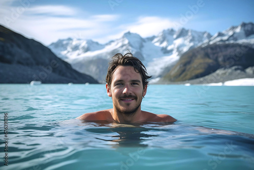 A handsome man in a swimming pool looks into the camera against a backdrop of majestic mountains and glaciers creating a serene atmosphere, Winter Retreat, winter recreational, winter swimming © Kate Simon