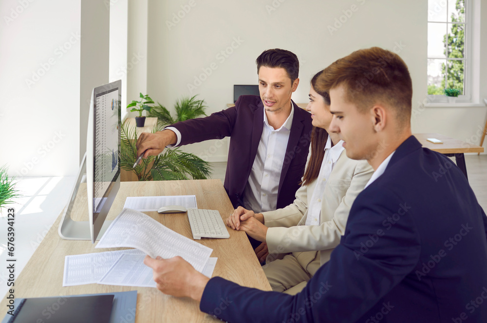 Young business team having work meeting in office. Group of three people sitting by office desk with modern computer. Male manager shows data charts on screen and explains some financial figures