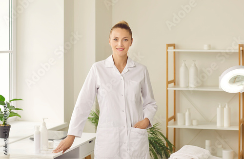 Portrait of professional beautician at work. Happy female cosmetologist in her workplace. Beautiful young woman wearing white coat standing by table with beauty products, looking at camera and smiling
