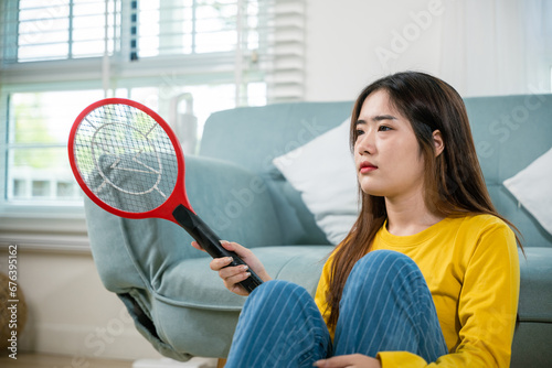Asian female sitting floor using mosquito device swatter or electric net racket, Young woman killing mosquitoes hand holding fly swatter like weapon in living room at home, technology insect killer photo