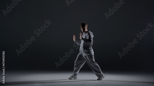 A young man stands against a dark background. He is highlighted by a white light from below. Creates a light haze. Demonstrates a dance movement by extending his arms forward. He is plastic, rhythmic