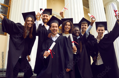 Group of happy smiling beautiful diverse multiethnic students in black graduation caps and gowns holding diplomas and standing together outside university building, with white columns in background