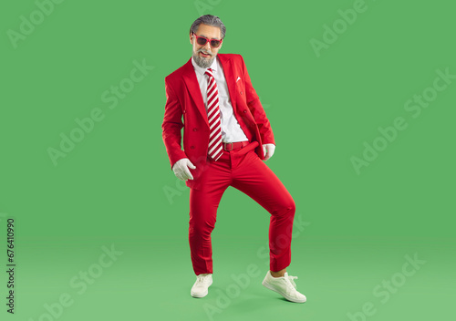 Senior playful grey haired showman in red sunglasses and red suit, white gloves, shirt and red tie posing on green background. Banner for Christmas, Xmas, New Year advertisement, sales, offer concept.