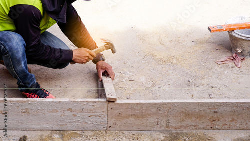 Construction builder worker uses a hammer to driving iron nail into a wooden beam at building site for guideline frame to filling formwork with wet concrete to build concrete floor on the work area.