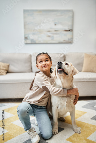little cute elementary age girl smiling and hugging labrador in modern apartment, pet and child