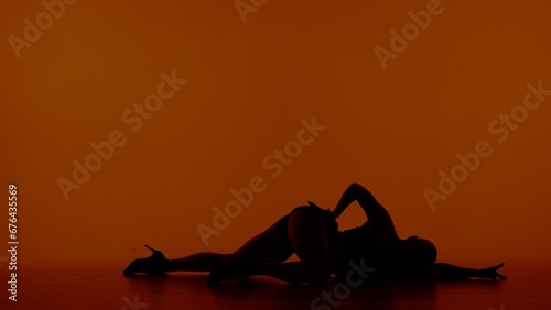 In the frame on a red orange background. Woman in silhouette on her open clothes and high heels. Demonstrates dance movement, pose. Shes lying on the floor with her hip up. Shes playing sexy, plastic