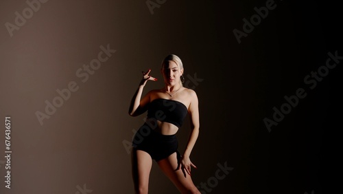 In the shot, a close up on a gray, darkened background. Woman stands on her a shadow falls. Dressed in open clothes. Shows a dance move, poses, hands up and looks into the camera. Its sexy, plastic © kinomaster