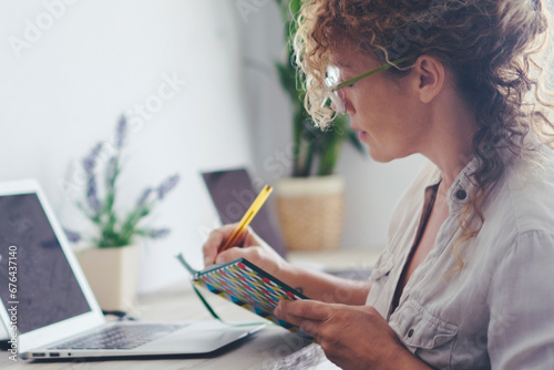 Modern housewife manage family economy at home - single lady use laptop computer and book with pen to takes notes and work - adult female viewed from side sitting and working at the desktop