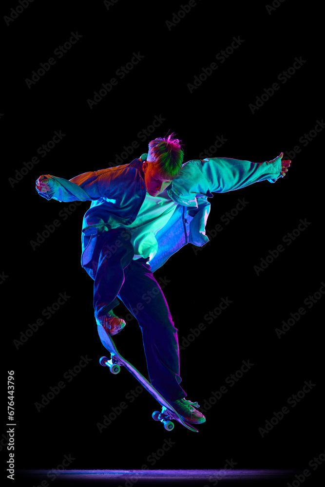 Teen boy, active guy in casual clothes in motion, training, practicing stunts on skateboard against black studio background in neon light. Concept of professional sport, competition, training, action