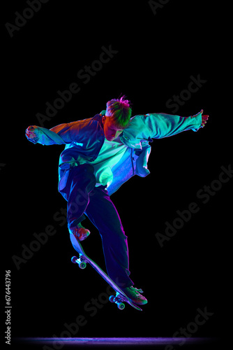 Teen boy, active guy in casual clothes in motion, training, practicing stunts on skateboard against black studio background in neon light. Concept of professional sport, competition, training, action