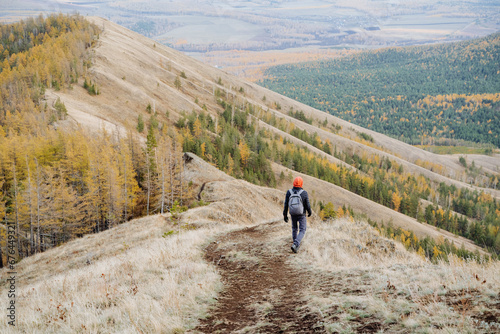 A hiker with a backpack walks alone along a mountain trail. Autumn time in hilly foothills, Rear view of man walking down the mountain.