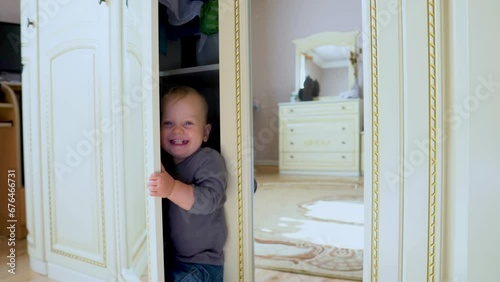 Joyful Caucasian baby with blond hair and blue eyes. Happy funny little boy playing hide and seek at home in a closet room. Child at home. Happy family