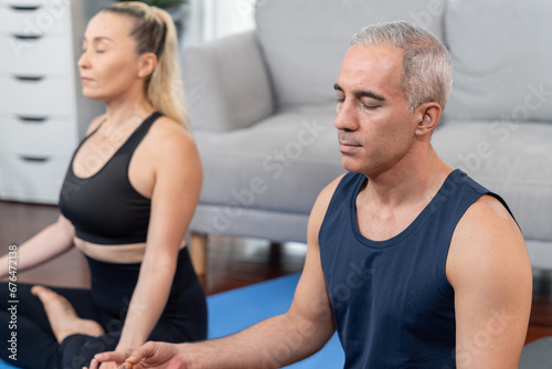 Happy active senior couple in sportswear being doing yoga in meditation posture on exercising mat at home. Healthy senior pensioner lifestyle with peaceful mind and serenity. Clout