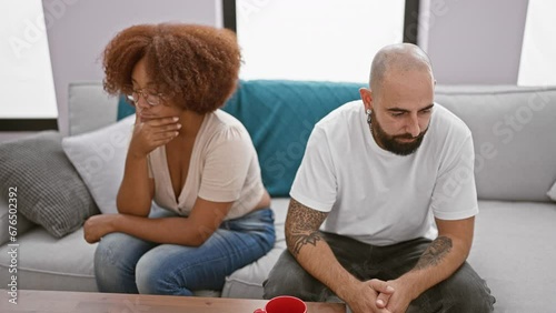 Beautiful interracial couple sitting in silence on living room sofa, stress and anger visible in expression, undercurrent of disagreement at home signifies relationship troubles photo