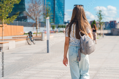 A beautiful young girl in jeans with long hair with a backpack walks down the street with a view of a high-rise building in St. Petersburg