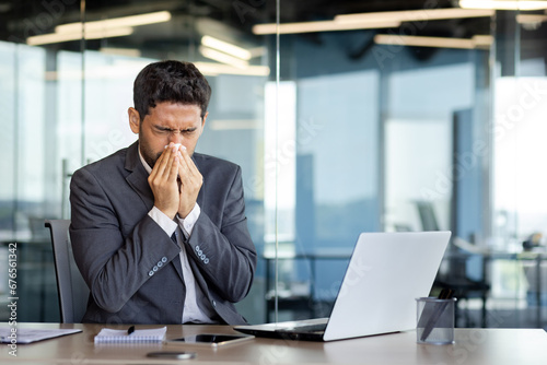 A man sneezes into a tissue at a workplace inside an office, a businessman is sick with a runny nose, works at a workplace with allergies, uses a laptop at work. photo