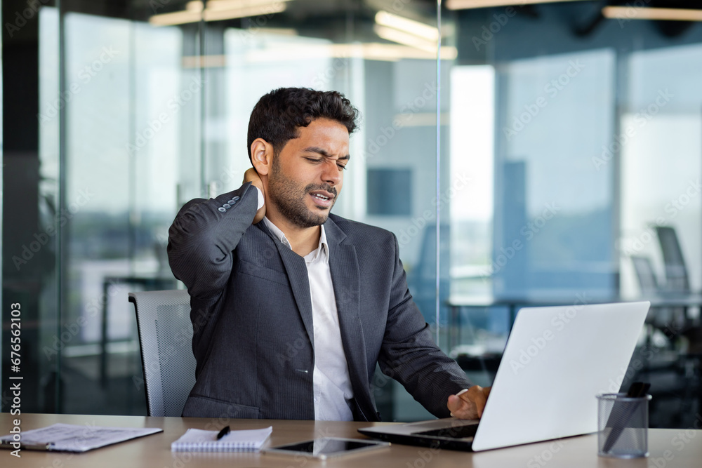 Neck pain, overworked and overtired man working late, businessman massaging neck muscle, working sitting on chair at table with laptop inside office at workplace. Neck pain, overworked and overtired man working late, businessman massaging neck muscle, working sitting on chair at table with laptop inside office at workplace.