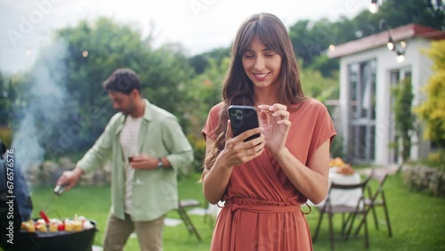 Portrait of young beautiful woman holding smartphone, smiling, texting, chatting and looking at phone screen. Leisure, holidays, eating, people and food concept - friends having party at house yard.
