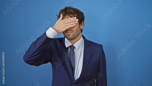 Sightless young hispanic man with a standoffish expression, hiding his sadness behind a hand covering his eyes. poignant denial concept amid blue isolated background. photo