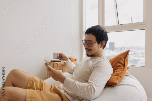 Happy asian man in long sleeve sit relaxed on bean bag using smartphone.