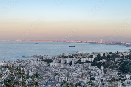 Algiers city aerial view from Bologhine mountain with ships anchored on the sea bay and the Great mosque minaret enlightened with golden hour sunlights. Admiralty lighthouse and Babeloued district.