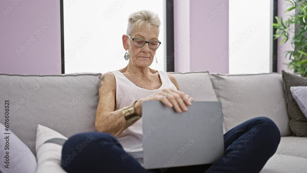 Confident and happy, a senior grey-haired woman with glasses, sitting on a comfortable sofa at home, is closing her laptop with a joyous smile after relaxing online work. Confident and happy, a senior grey-haired woman with glasses, sitting on a comfortable sofa at home, is closing her laptop with a joyous smile after relaxing online work.